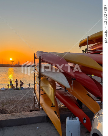 Evening beach scene with stacked kayaks in the foreground and a family enjoying the water during sunset. The golden sky creates a calm atmosphere as they play by the shore. 128327897