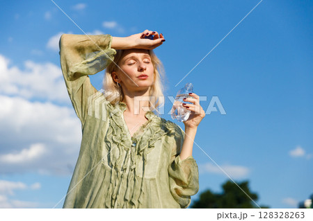A Young Woman Relaxes Outdoors, Overcoming the Summer Heat While Holding a Water Bottle and Taking a Moment to Enjoy the Beautiful Blue Sky and Fluffy Clouds Above 128328263