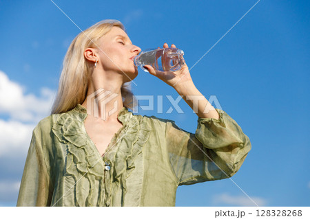 Refreshing Hydration: A Woman Enjoying Water Under a Bright Blue Sky in a Soft Green Blouse with Light Ruffled Details in Nature's Embrace 128328268