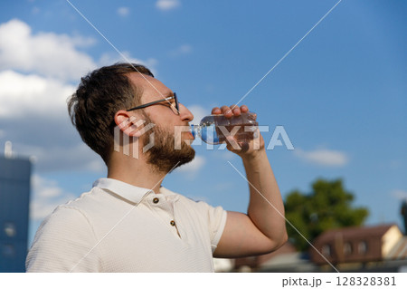 A Man Quenching His Thirst with a Bottle of Water Under a Bright Blue Sky with Fluffy Clouds in the Background in an Outdoor Setting A Man Quenching His Thirst with a Bottle of Water Under a Bright Blue Sky with Fluffy Clouds in the Background in an Outdoor Setting 128328381