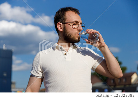 Man Quenching Thirst: Stylish Individual Enjoying Refreshing Bottled Water Under Clear Blue Sky on a Bright Day with Cloudy Background in Urban Setting Man Quenching Thirst: Stylish Individual Enjoying Refreshing Bottled Water Under Clear Blue Sky on a Bright Day with Cloudy Background in Urban Setting 128328385