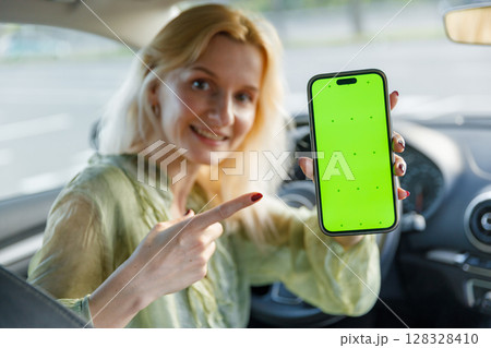 A Smiling Woman Inside a Car Holding a Smartphone with a Bright Green Screen, Engagingly Pointing at It to Showcase the Device's Unique Features and Vibrant Display 128328410