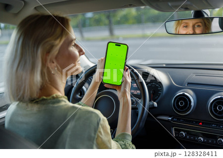 A Woman in a Car Holding a Smartphone with a Bright Green Screen While Driving, Showcasing Modern Technology Usage and Driver Engagement in Daily Commute Activities A Woman in a Car Holding a Smartphone with a Bright Green Screen While Driving, Showcasing Modern Technology Usage and Driver Engagement in Daily Commute Activities 128328411