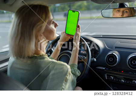 Young woman inside a car holding a smartphone with a bright green screen while driving, showcasing modern technology usage during travel or navigation. 128328412