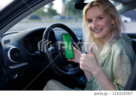 Happy Young Woman Displaying a Smartphone While Sitting in a Car and Showing a Thumbs Up Gesture with a Bright Smile, Emphasizing the Joy of Connectivity and Modern Technology 128328567