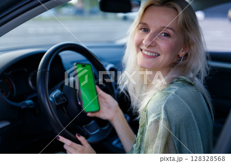 A Young Woman in a Car Holding a Smartphone with a Green Screen, Smiling Cheerfully, Capturing the Joy of Modern Technology and Life 128328568