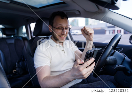 Excited Young Man Inside Car Celebrating Success on Smartphone After Receiving Good News or Winning a Game, Dynamic Expression of Joy and Achievement in His Posture, Inside Vehicle 128328582