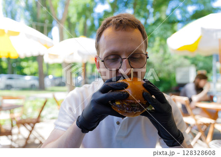 A Man with Glasses in a Caf Holding a Delicious Burger Carefully with Black Gloves, Enjoying a Bright Sunny Day Under Attractive Awning Shades. 128328608