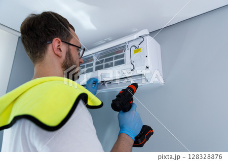 Professional technician performs routine maintenance on a modern air conditioning unit mounted on a wall, ensuring optimal performance in a comfortable indoor environment. 128328876