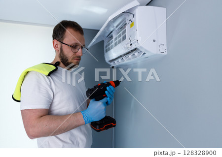 Man Performing Air Conditioner Installation or Repairs with a Power Drill and Wearing Protective Gear on His Hands and Shoulders for Safety and Hygiene in a Modern Indoor Environment 128328900