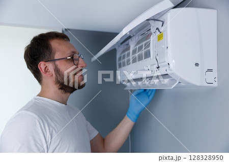 Man with Glasses Performing Maintenance on Air Conditioning Unit While Smiling and Wearing Blue Gloves in a Modern Room 128328950