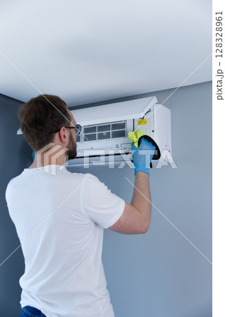A Person Cleaning an Air Conditioner Unit Mounted on a Wall in a Modern Room with Gray Painted Walls and a Bright Interior Design for Enhanced Air Quality and Efficiency 128328961
