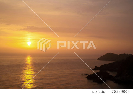 Spectacular Sunset Over Calm Waters: A Breathtaking View of the Sun Setting on the Horizon at Dusk Behind Serene Ocean Waves and Rocky Shorelines 128329009