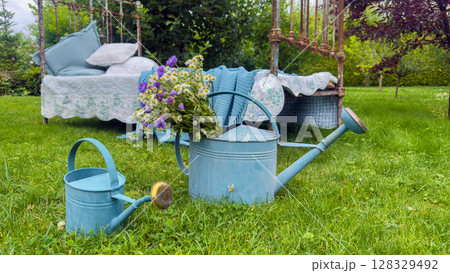 Charming Garden Scene Featuring Colorful Watering Cans and Floral Arrangement Set Against a Rustic Outdoor Bed in a Lush Green Lawn 128329492