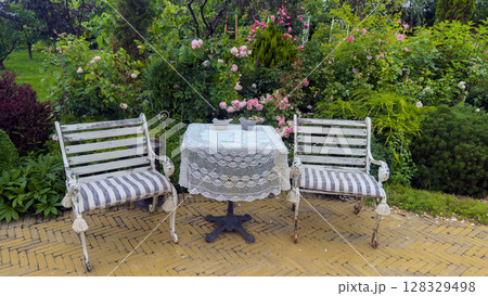 Charming Garden Setting with Rustic White Benches and a Lace-Covered Table Surrounded by Lush Roses and Greenery Creating a Serene Outdoor Retreat for Relaxation Charming Garden Setting with Rustic White Benches and a Lace-Covered Table Surrounded by Lush Roses and Greenery Creating a Serene Outdoor Retreat for Relaxation 128329498