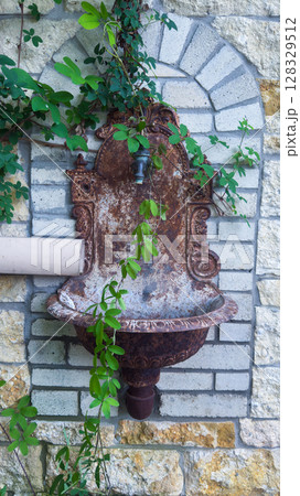 Rustic Wall Fountain Surrounded by Lush Green Vines Displaying a Charming Contrast Against the Textured Stone Wall in a Serene Outdoor Setting Rustic Wall Fountain Surrounded by Lush Green Vines Displaying a Charming Contrast Against the Textured Stone Wall in a Serene Outdoor Setting 128329512