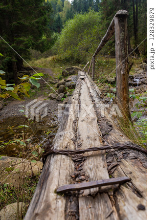 A Serene Woodland Path Featuring a Weathered Wooden Bridge Over a Gentle Stream Surrounded by Lush Green Foliage and Towering Trees in an Idyllic Natural Setting 128329879