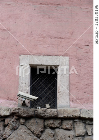 Surveillance Camera Overlooking a Shaded Window Set Against a Pink Wall with Stone Base: A Blend of Modern Security and Historical Architecture's Repose Surveillance Camera Overlooking a Shaded Window Set Against a Pink Wall with Stone Base: A Blend of Modern Security and Historical Architecture's Repose 128330196