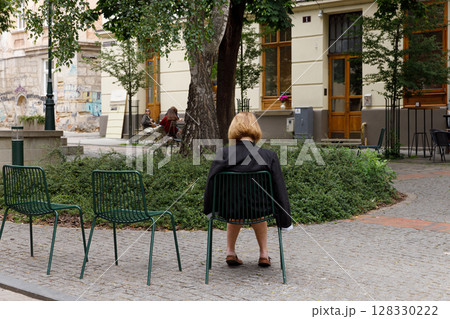 A Serene Outdoor Scene Featuring a Woman Sitting Back on a Chair in a Quaint European Street Surrounded by Lush Greenery and Architectural Beauty 128330222