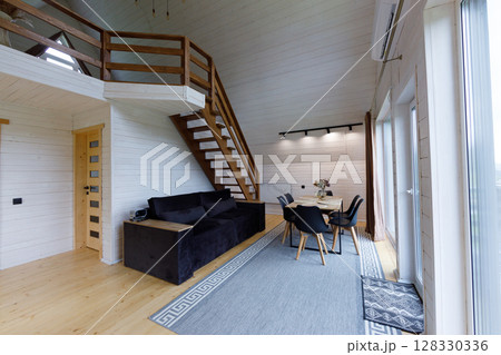 Cozy Modern Interior of a Wooden Loft with Open Space Featuring Stylish Staircase, Comfortable Couch, Dining Table and Natural Light Streaming Through Large Windows 128330336