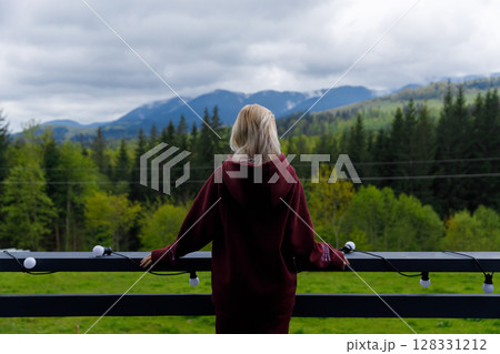 A Serene Moment of Contemplation: A Young Woman Enjoys Nature's Beauty from a Scenic Balcony Overlooking Majestic Mountains and Lush Green Forests in a Peaceful Setting 128331212