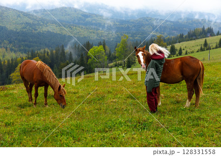 A Serene Moment in Nature: A Young Individual Interacting with Two Horses Surrounded by Lush Green Mountains and Scenic Wilderness Landscape A Serene Moment in Nature: A Young Individual Interacting with Two Horses Surrounded by Lush Green Mountains and Scenic Wilderness Landscape 128331558