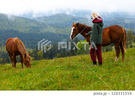 A Serene Moment: A Girl Interacting with Horses in a Picturesque Mountain Landscape Surrounded by Lush Grass and Scenic Beauty, Perfect for Nature Lovers and Equestrian Enthusiasts 128331559