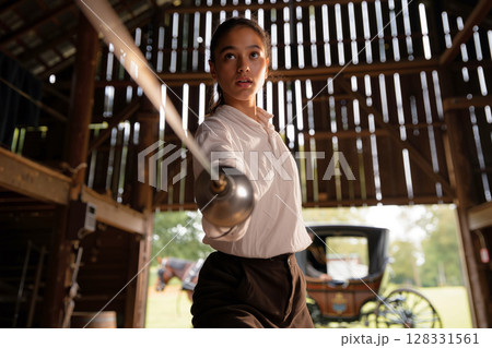 Young Woman Practices Sword Fighting in Rustic Barn Setting Surrounded by Horse and Carriage 128331561