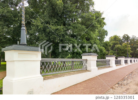 Park fence made of forged metal lattice with stone stobles, against the background of green trees 128335301