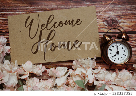Welcome Home text with alarm clock and flowers decoration on wooden background Welcome Home text with alarm clock and flowers decoration on wooden background 128337353