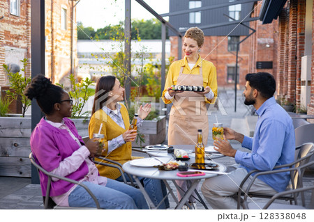 A group of friends enjoying sushi and drinks at an outdoor restaurant, served by a friendly waitress. A group of friends enjoying sushi and drinks at an outdoor restaurant, served by a friendly waitress. 128338048