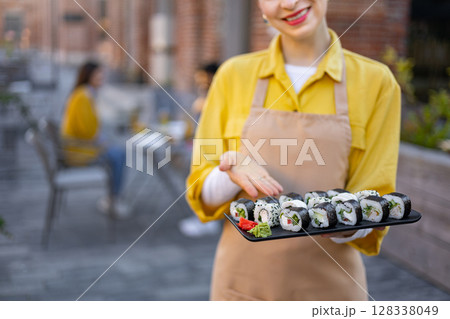 A server presents a platter of sushi rolls at an outdoor restaurant, smiling with pride. Guests can be seen in the background. 128338049