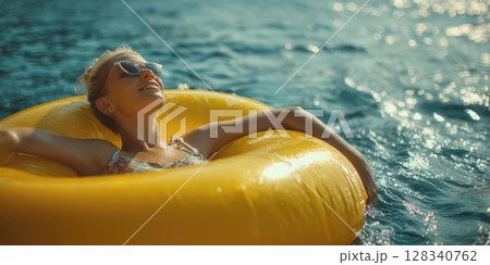 A beautiful woman, eyes closed in contentment, soaks up sun while floating effortlessly on yellow inflatable ring in sea water. Ideal for themes of summer relaxation, vacation, serene aquatic leisure 128340762