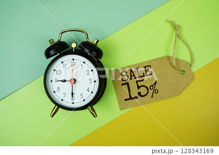 Sale 15% text on price tag with alarm clock and trolley shopping cart on green, yellow geometric background 128343169