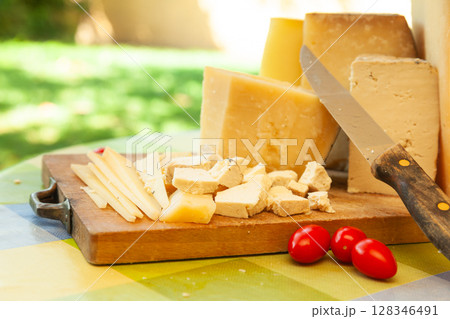 Assorted blocks and slices of hard cheese on wooden board with knife and tomatoes in outdoor summer setting 128346491