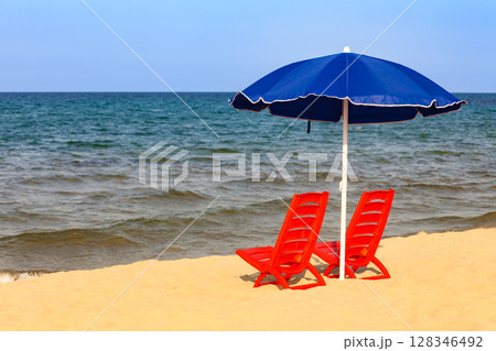 Red beach loungers and blue umbrella set on sandy shore with sea view Red beach loungers and blue umbrella set on sandy shore with sea view 128346492
