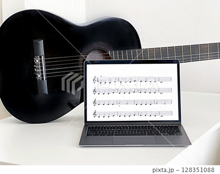 A black acoustic guitar rests beside a laptop displaying musical notes on sheet music.  128351088