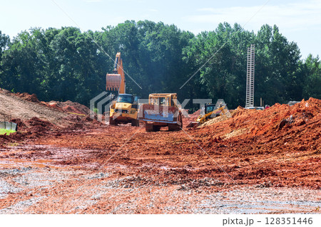 Heavy machinery navigates muddy construction site surrounded by trees, showing ongoing development activity. Heavy machinery navigates muddy construction site surrounded by trees, showing ongoing development activity. 128351446