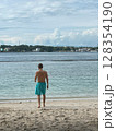 Man in swim shorts on sandy beach, looking at tranquil ocean. 128354190