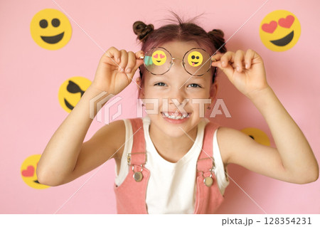 A beautiful girl on a pink studio background with a variety of emoticons pasted on glasses. World Social Media Day. Smile icons and emojis 128354231