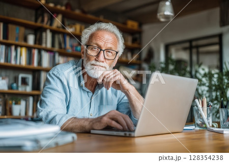 Thoughtful elderly man with glasses working on a laptop in a cozy home office filled with books and Thoughtful elderly man with glasses working on a laptop in a cozy home office filled with books and 128354238