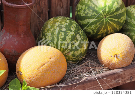 Close view of fresh ripe melons and watermelons in a wooden cart with straw. Bright rustic composition, ideal for farmers market promotion or packaging design. 128354381