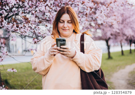 Happy 30s woman cheerfully talking on a smartphone against a background of pink tree blossoms. Spring time in the city. 128354879