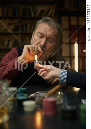 A man lights a cigarette, another man offers assistance, while playing poker A man lights a cigarette, another man offers assistance, while playing poker 128355347