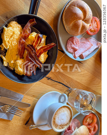 Delicious breakfast with scrambled eggs and crispy bacon served in a pan, alongside fresh bread, sliced meats, and tomato. Accompanied by a frothy cappuccino in a cozy morning setting. 128356367