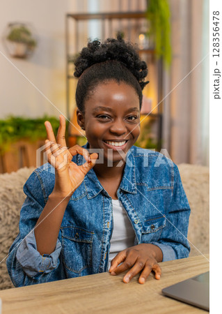 African American young woman smiles showing okay hand gesture while sitting at table on sofa at home 128356478