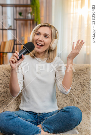 Adult woman singing with headphones and microphone at home dancing joyfully feeling like a superstar 128356482