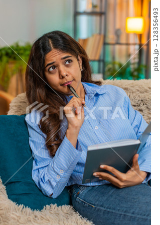 Indian woman writing thoughts in diary with gentle smile, reflecting with peaceful calm expression 128356493