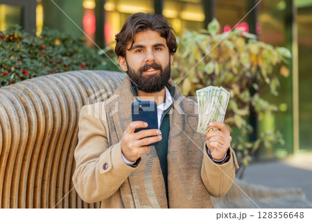 Happy rich young Indian businessman using smartphone and counting money on downtown city street Happy rich young Indian businessman using smartphone and counting money on downtown city street 128356648