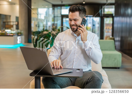 Businessman in formal shirt answering to client on mobile phone call, using laptop in office lobby 128356650